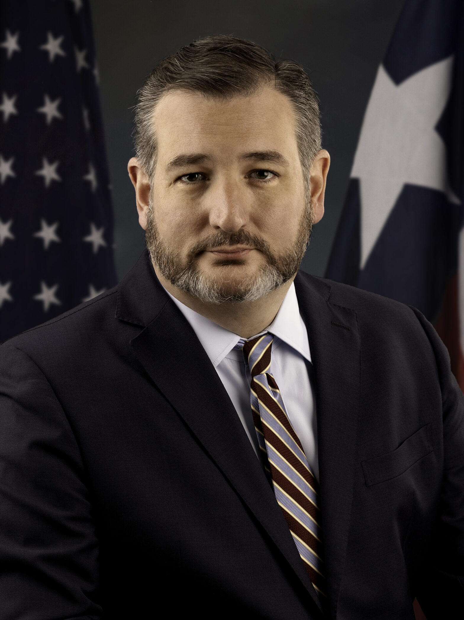 Head-and-shoulders portrait of a man in a dark suit and striped tie, with flags in the background.